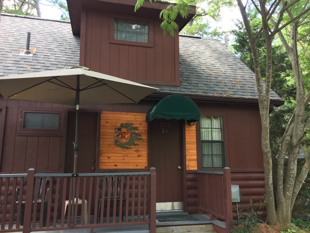 A small, rustic, brown cabin with a front porch and a deck with a black umbrella. A green awning is over the front door, with a wooden sign next to it.