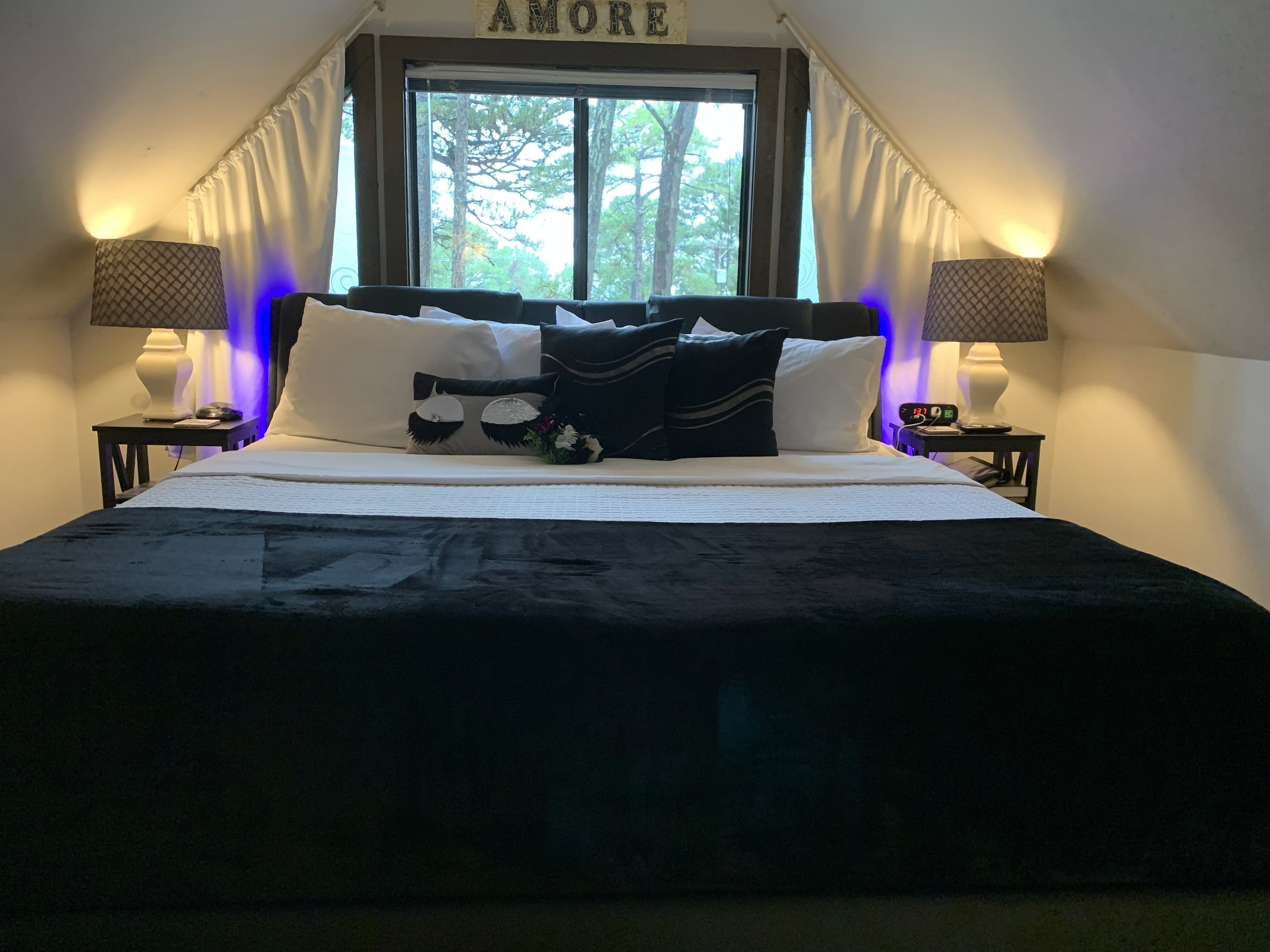 A loft bedroom features a king-size bed with black and white bedding. The room has low sloped ceilings, a window with white curtains, and a sign above the bed that says "AMORE."