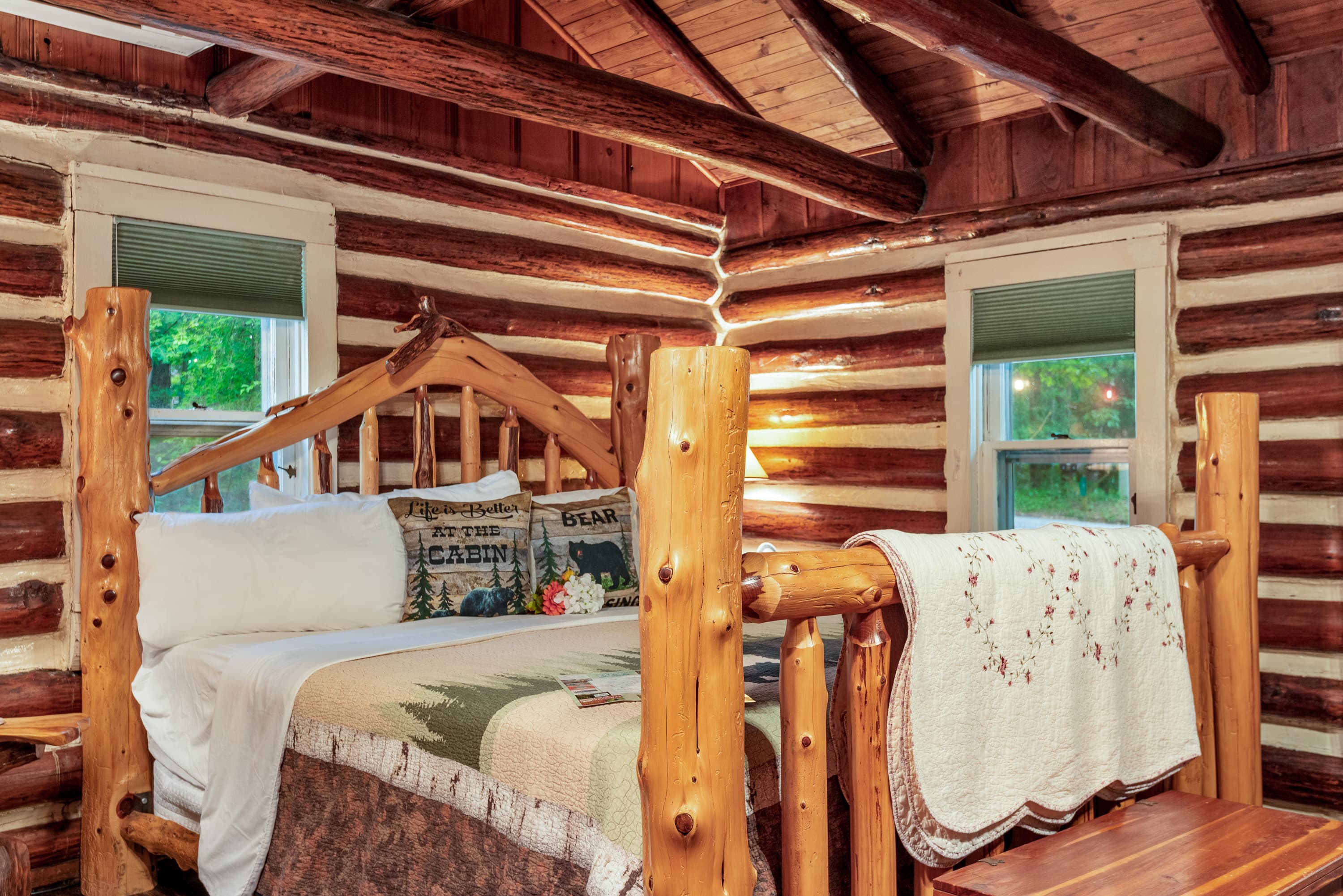 A rustic log cabin bedroom with a king bed featuring a decorative, wooden log headboard. The walls are also made of natural wood logs.