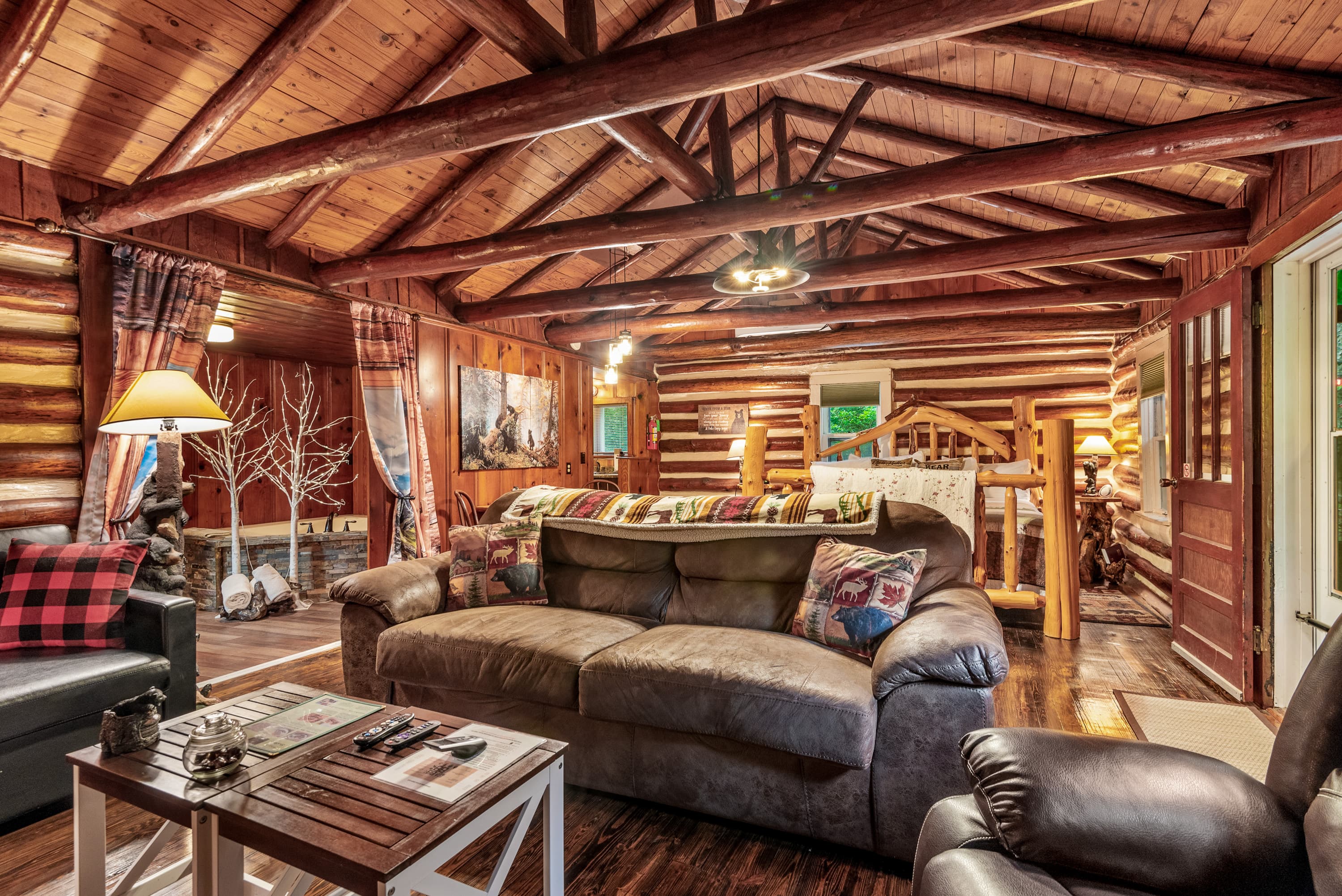 A spacious, rustic log cabin interior features a leather couch and armchair, a coffee table, and a small area rug with a king-size log bed in the background. The walls and ceiling are made of wood logs and beams.