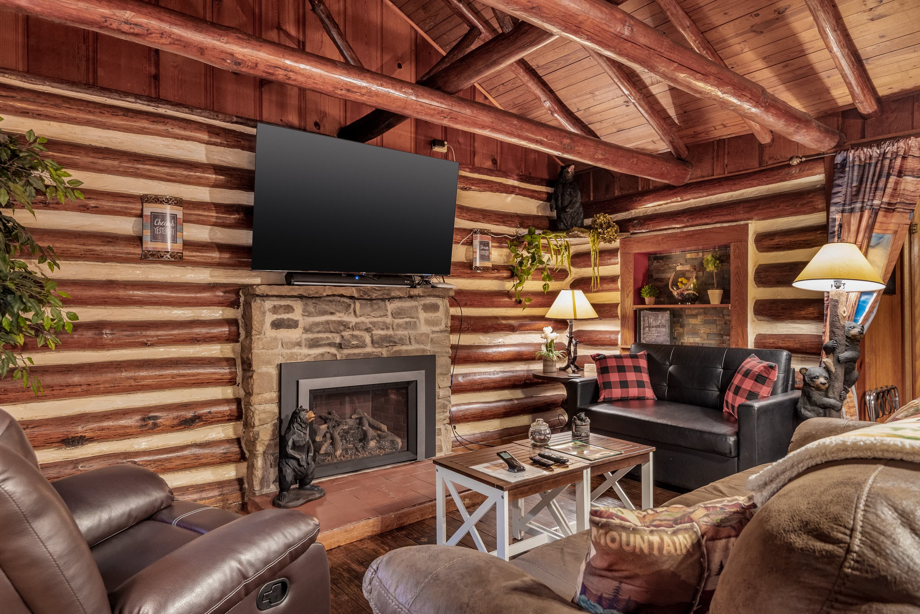 A rustic log cabin sitting area features a stone fireplace with a flat-screen TV mounted above it. The room has wood log walls, leather furniture, and a coffee table.