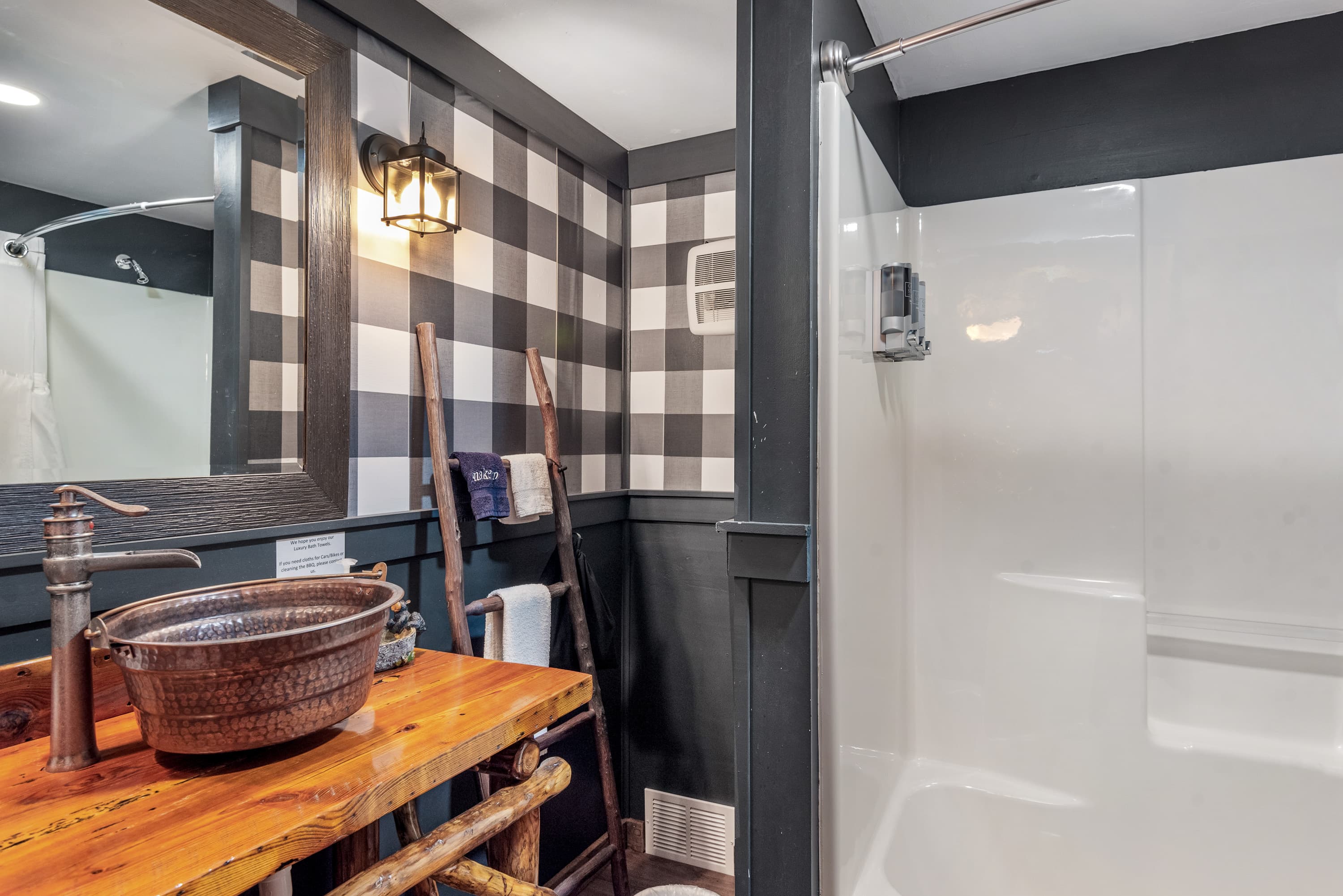 A modern rustic bathroom features black and white buffalo check wallpaper. The room has a wood vanity with a bowl sink and a shower with a white curtain.