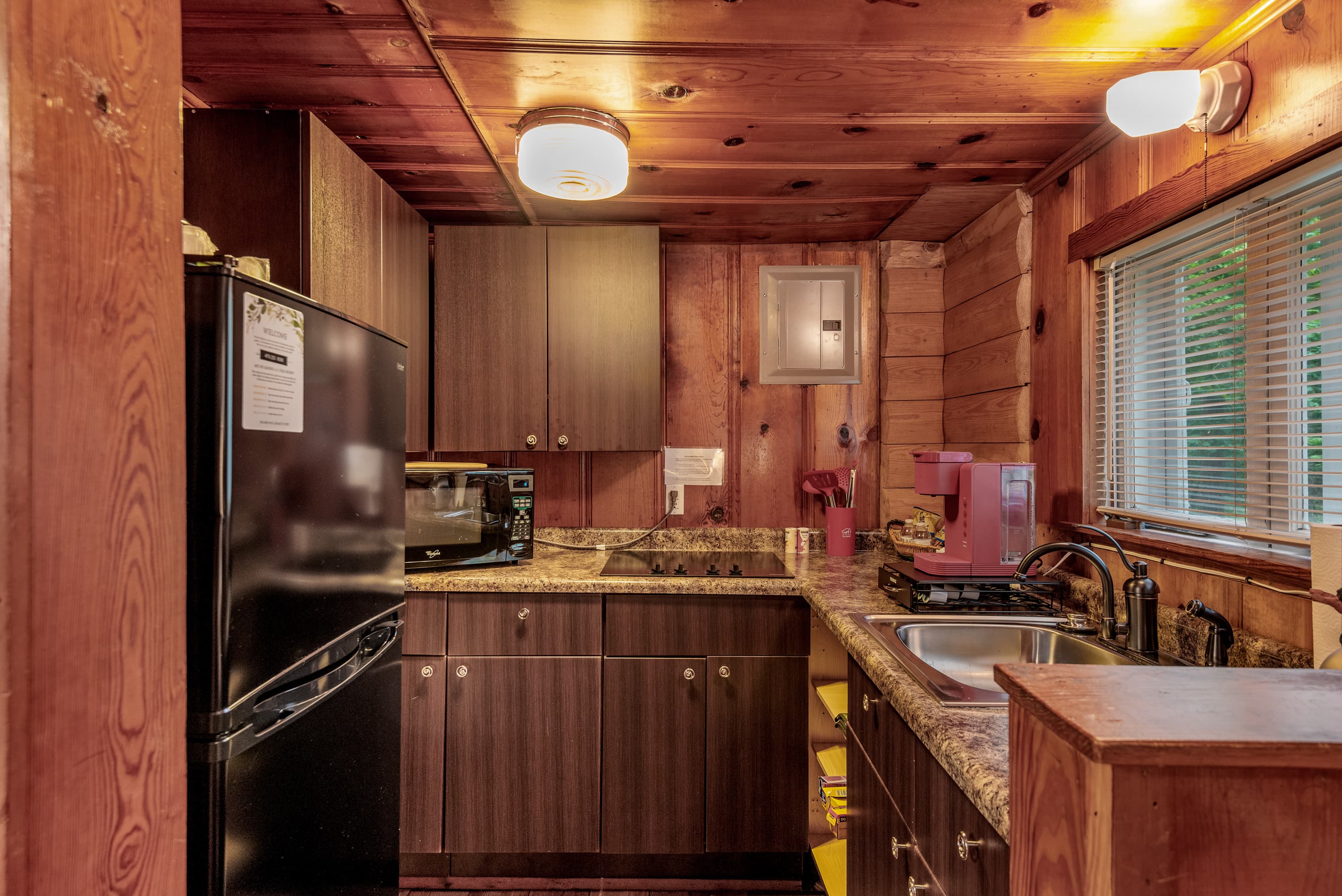 A rustic kitchen with wood-paneled walls and dark wood cabinets. The room features a refrigerator, a sink, and a stovetop.