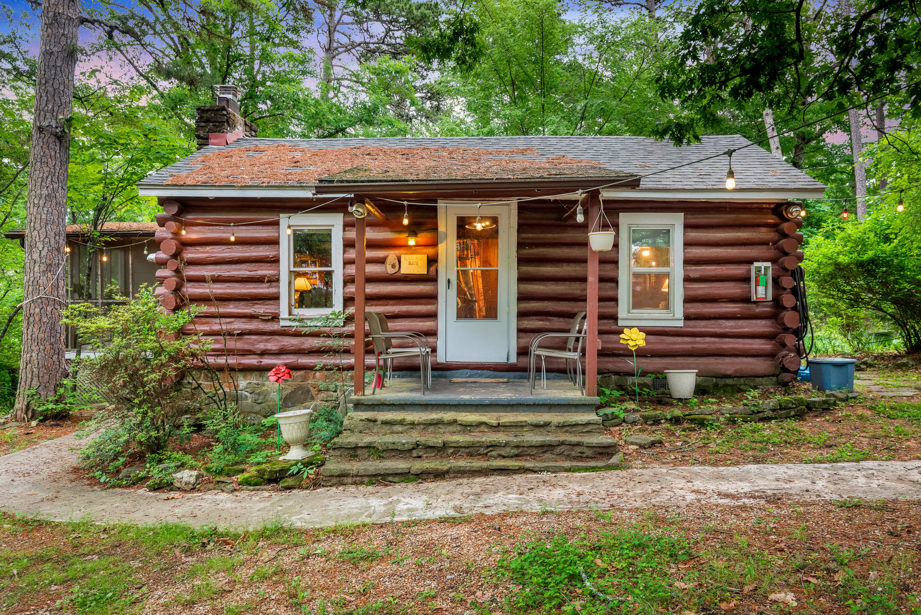 A cozy, rustic log cabin with a small porch and a wooden deck. The porch features a table and two chairs and is surrounded by a garden and tall pine trees.