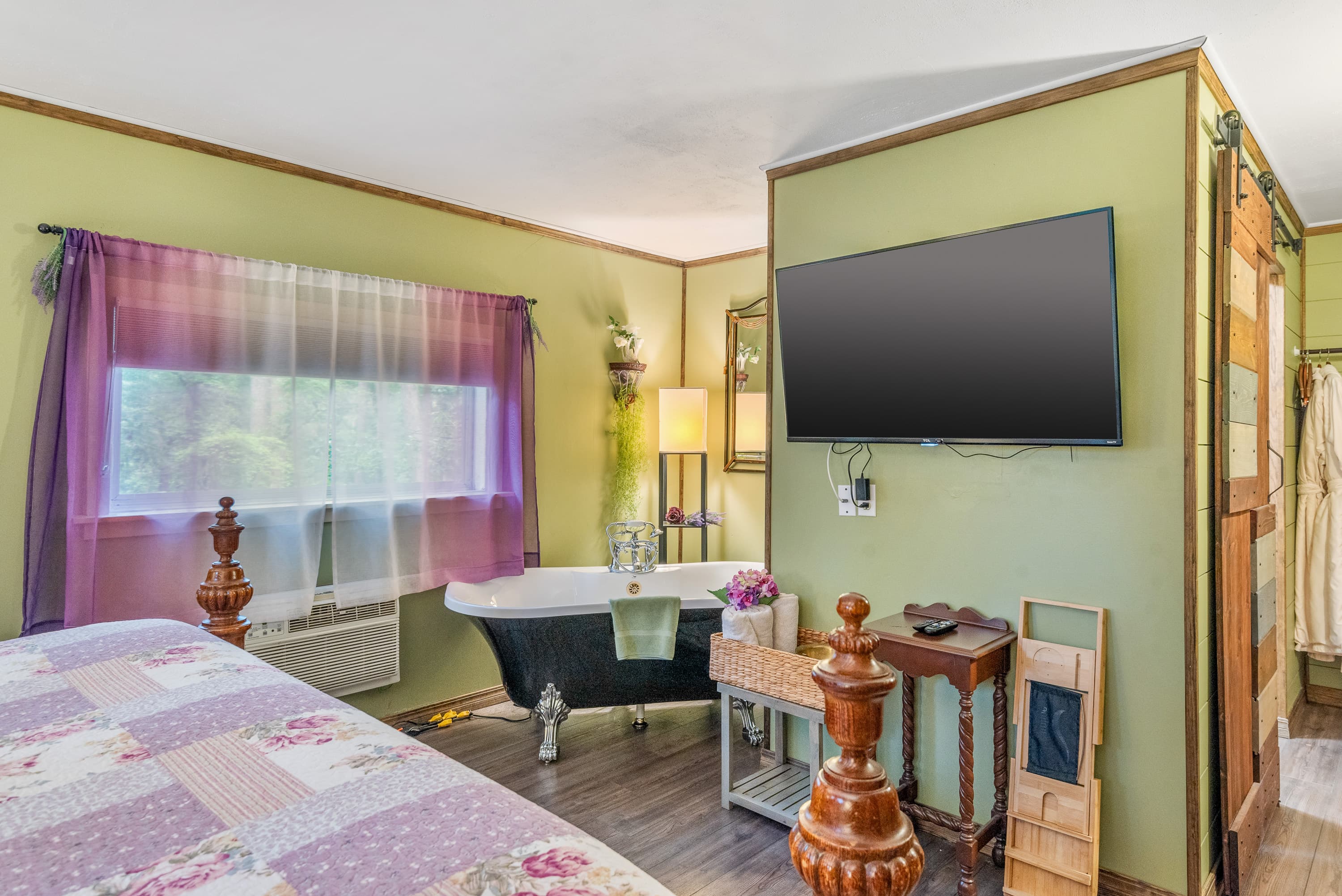 A cozy bedroom features a bed with a floral quilt, a large claw-foot tub, a TV mounted on a partial wall, and light green walls.