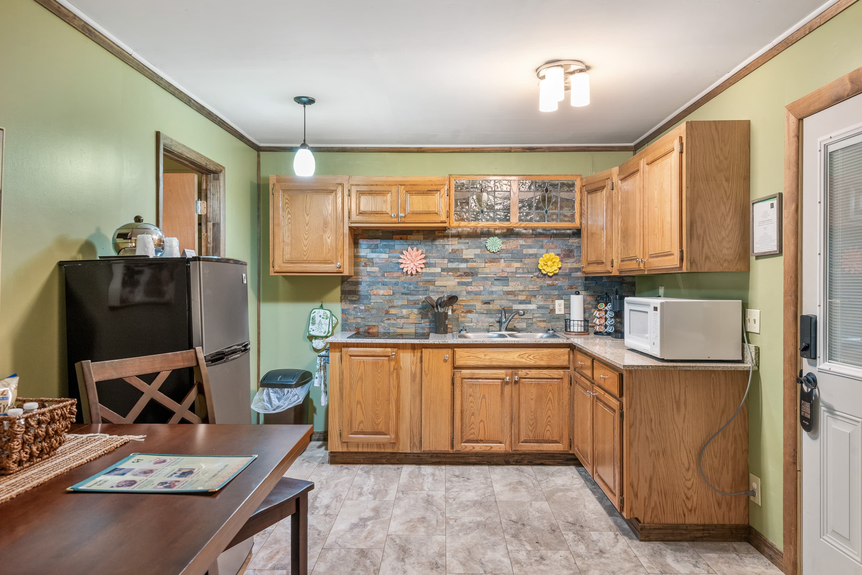 A rustic kitchen features wood cabinets and a stone backsplash. The room has light green walls, a small dining table, and a refrigerator.