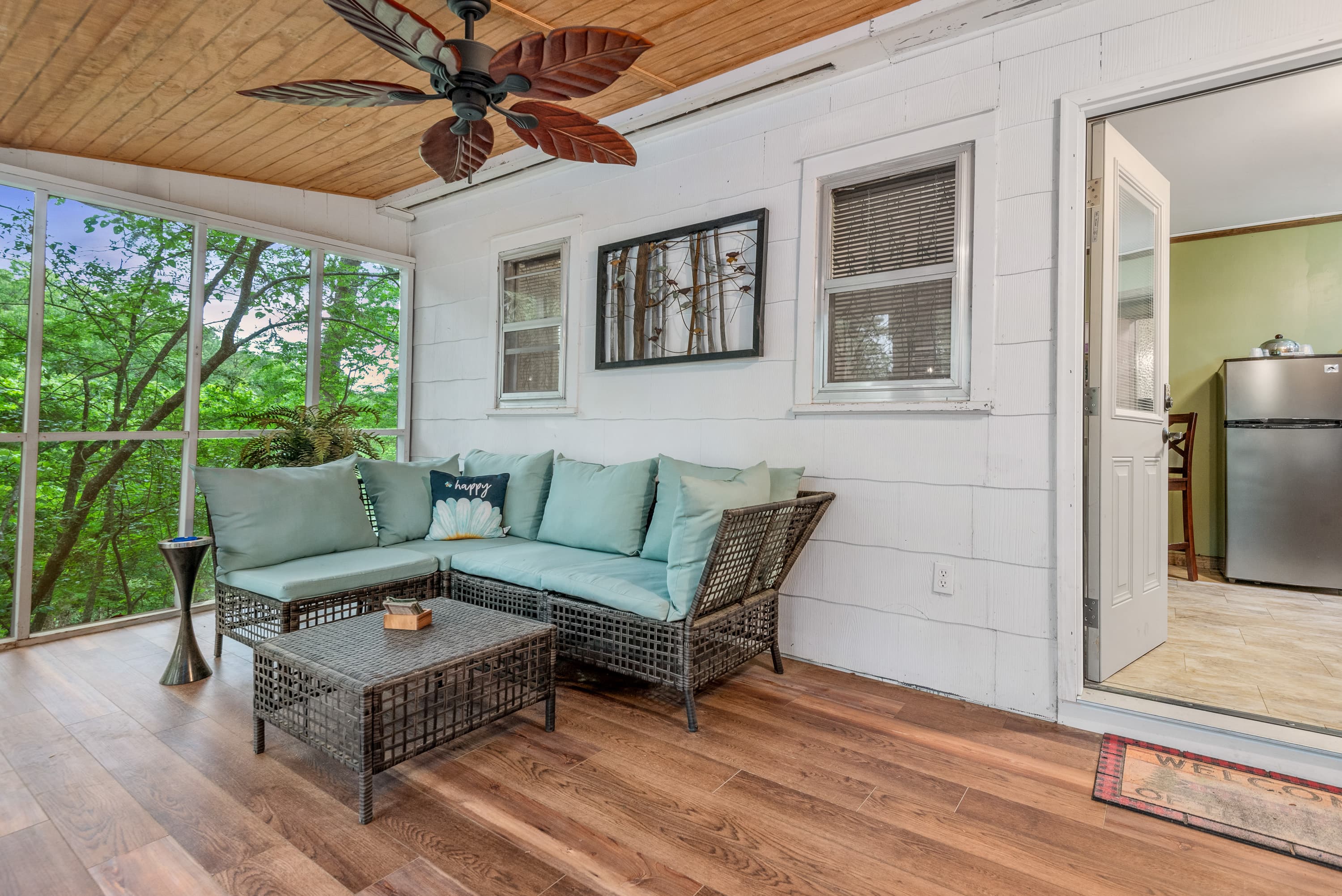 A comfortable screened-in porch with a wicker sectional couch, a coffee table, and a ceiling fan. The porch overlooks a wooded area.
