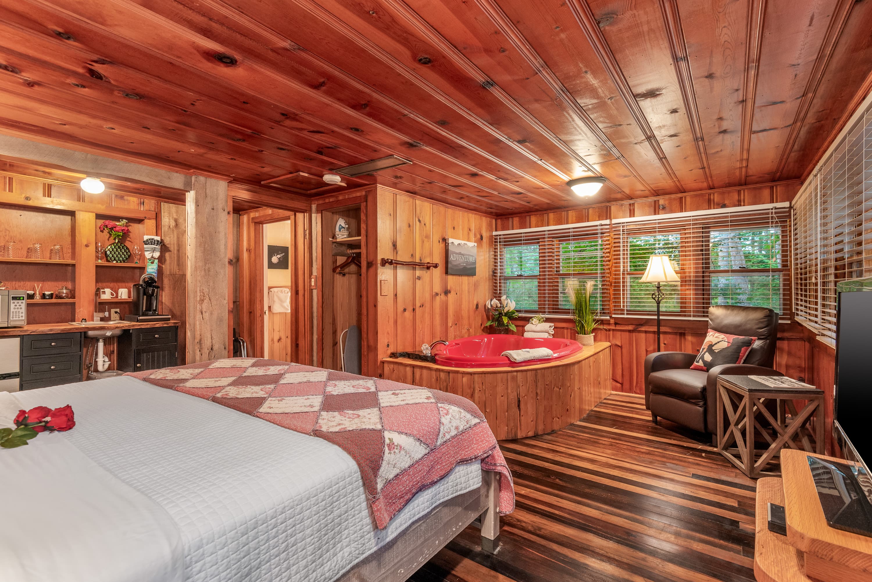 A spacious, rustic, wood-paneled bedroom with a king-size bed, a red jetted tub, and a comfortable leather armchair.
