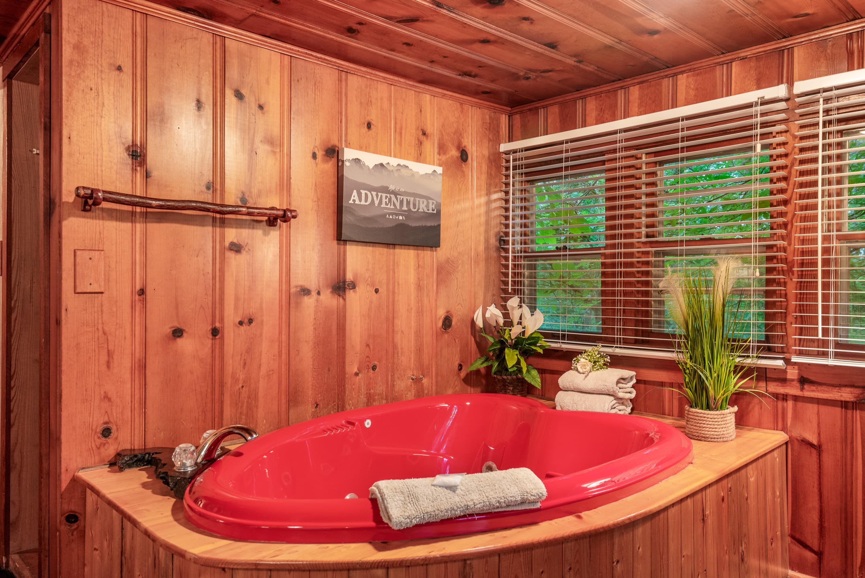 A red, heart-shaped jetted tub is in a rustic room with wood-paneled walls and blinds on the window.