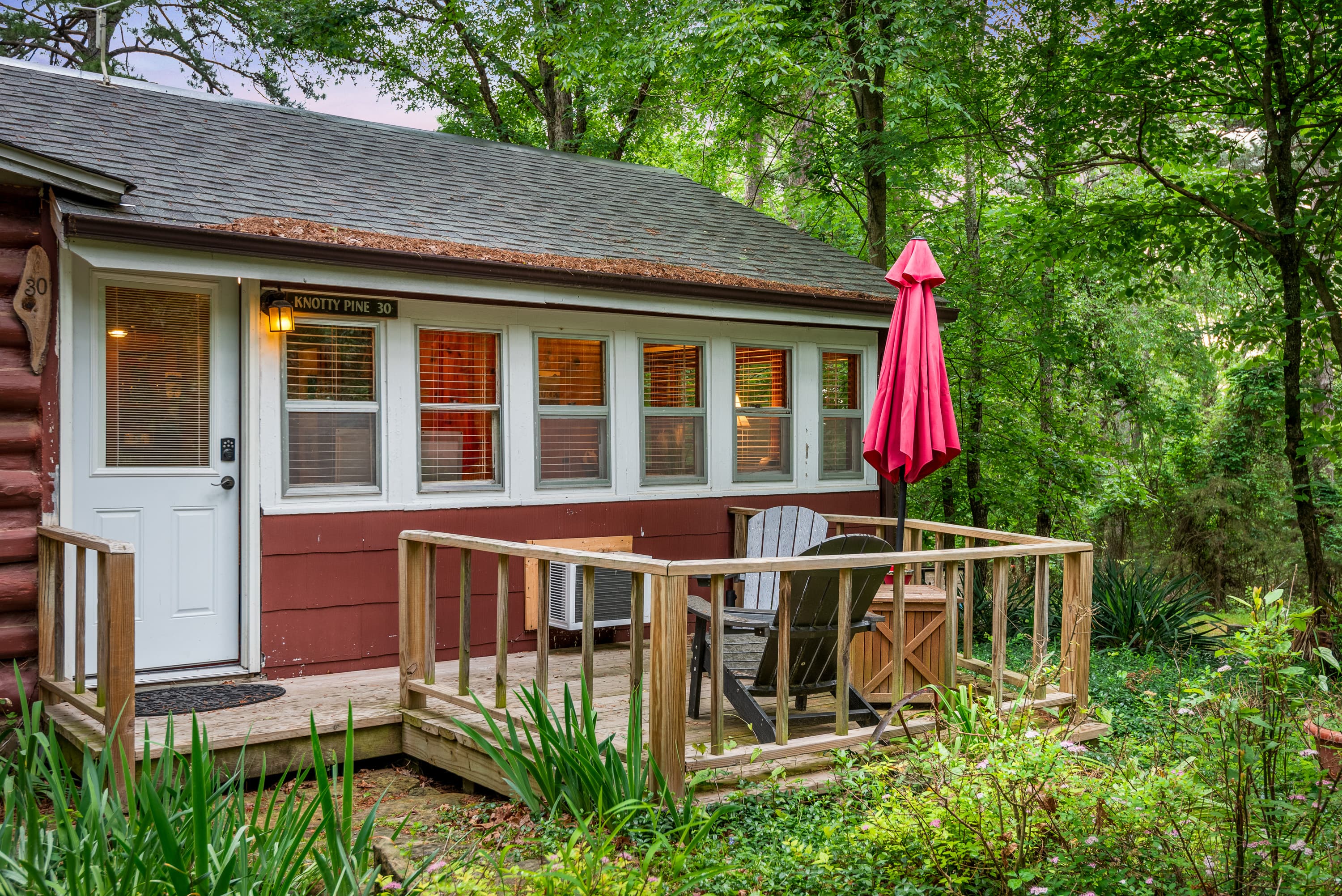 A charming, rustic cabin with a front porch and a wooden deck. The deck features a table, two chairs, and a red patio umbrella. The cabin is surrounded by a wooded area.