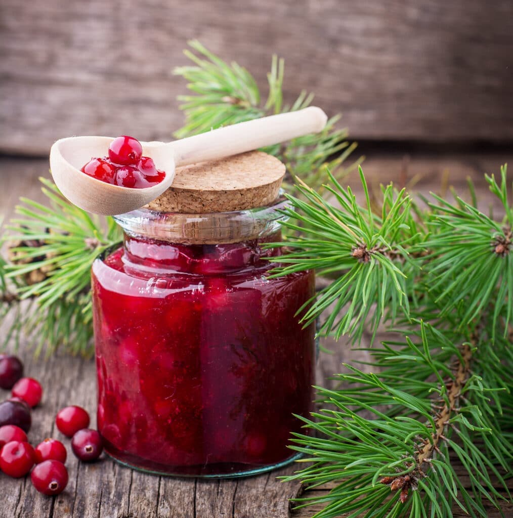 A jar of red jam topped with a wooden spoon sits beside pine branches on a wooden surface.