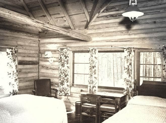Rustic log cabin interior featuring two beds, a table, and large windows with floral curtains.