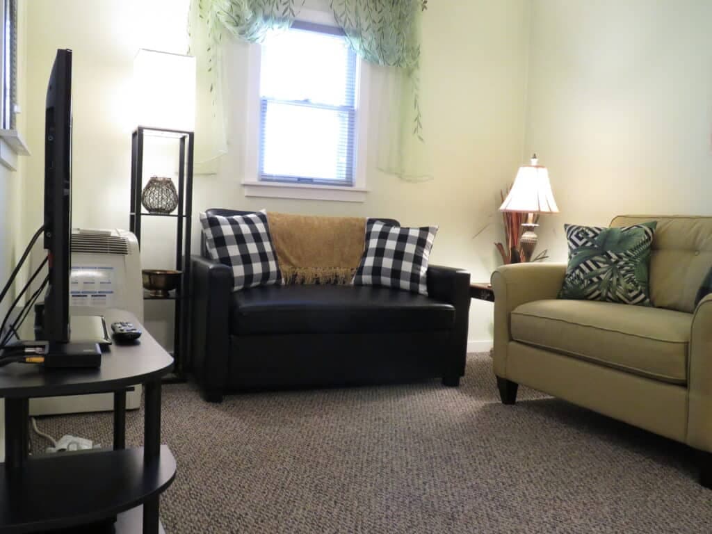 A cozy living room featuring a black couch with checkered pillows, a tan armchair, and a TV stand.