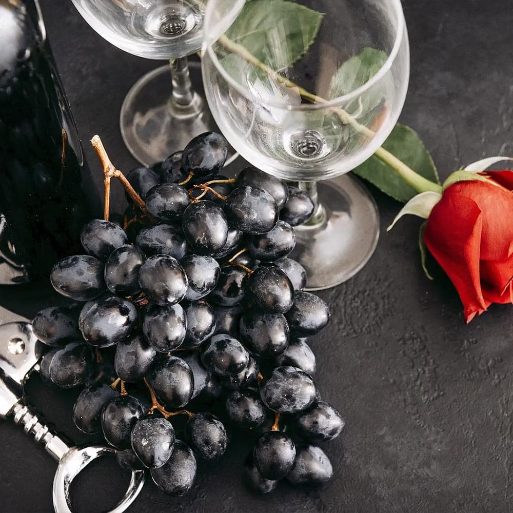 A cluster of black grapes beside two wine glasses and a red rose on a dark surface.