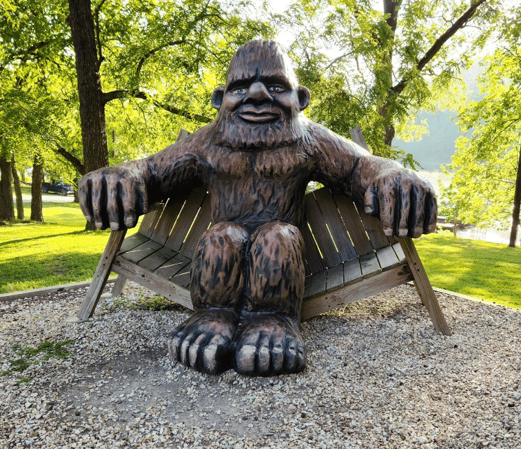 A large wooden sculpture of Bigfoot seated on a rustic bench surrounded by greenery.