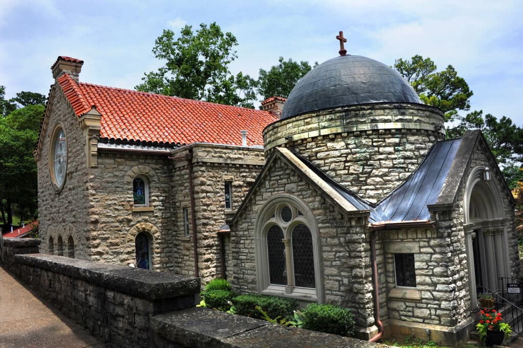A stone building with a domed roof and red-tiled accents surrounded by greenery.