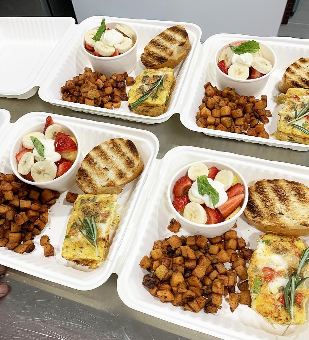 A neatly arranged breakfast featuring portions of scrambled eggs, grilled toast, seasoned potatoes, and fruit cups with strawberries and bananas.