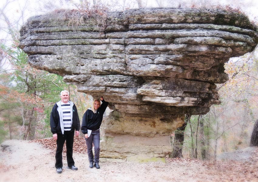 Two people stand beside a large, flat-topped rock formation in a wooded area.