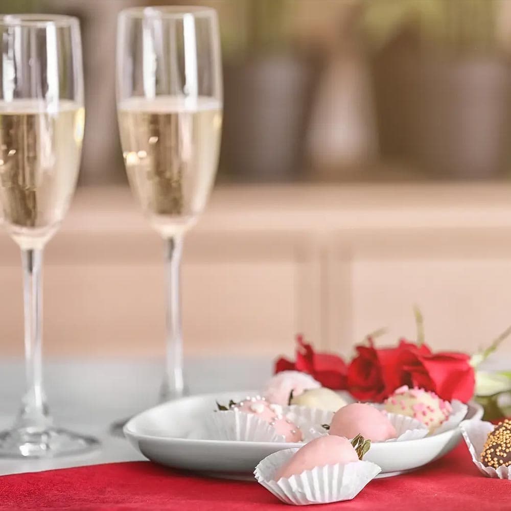 Two glasses of champagne next to a plate of decorated strawberries and red roses on a table.