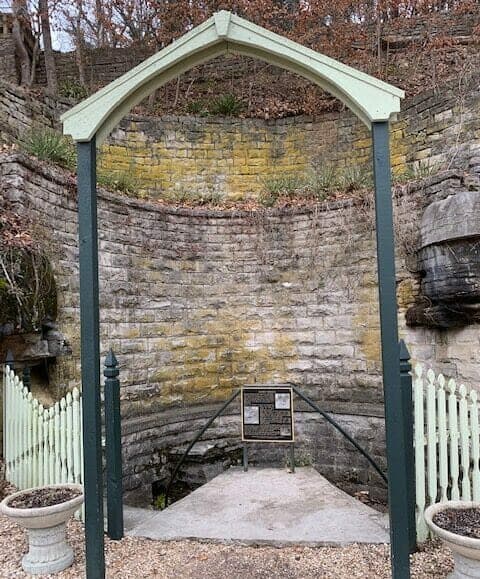 A rustic archway frames a stone structure with a plaque in a wooded area.