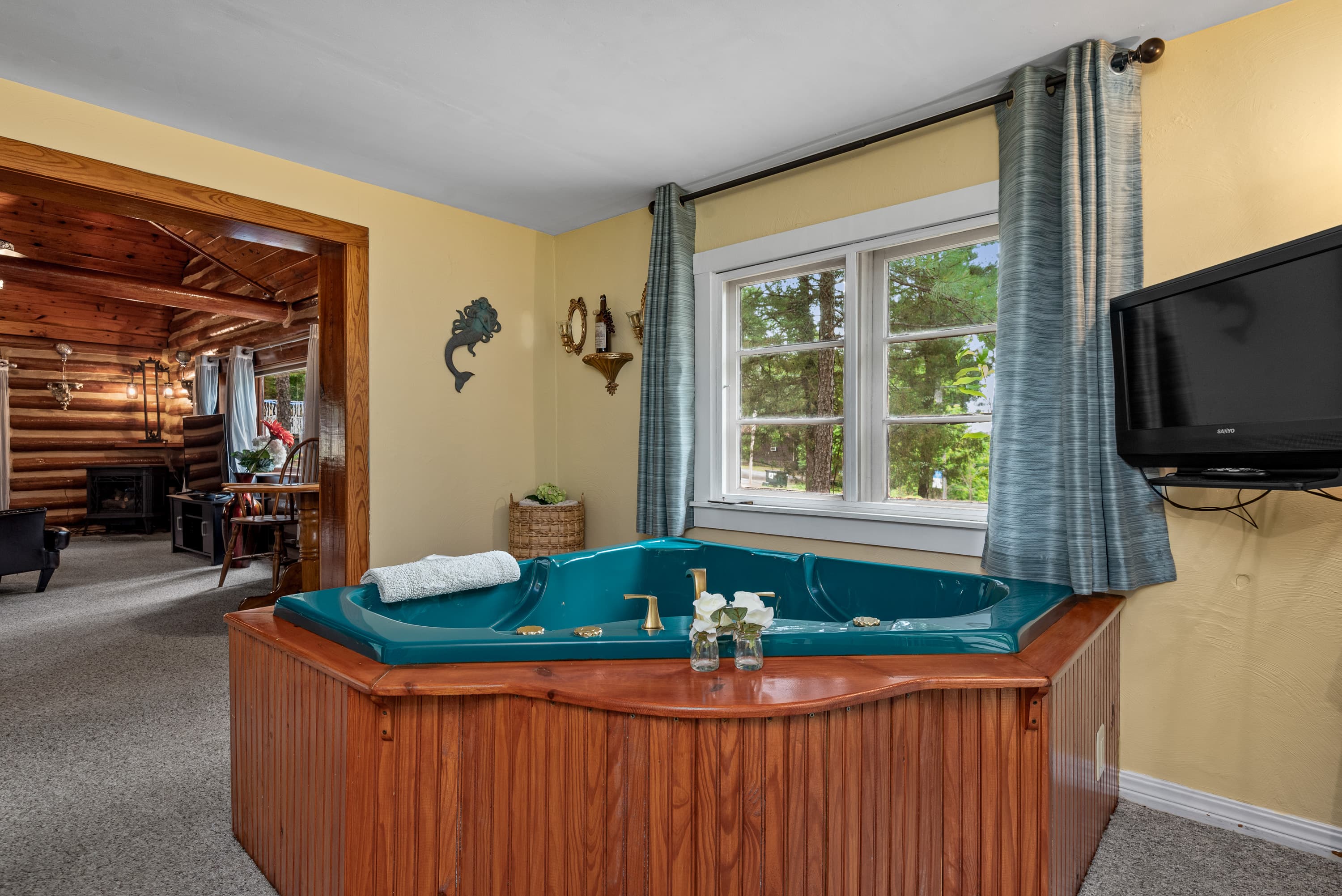 A large, turquoise, jetted tub with a wooden surround is in a rustic sunroom with yellow walls. A doorway on the left leads to a cozy log cabin living room with a fireplace.