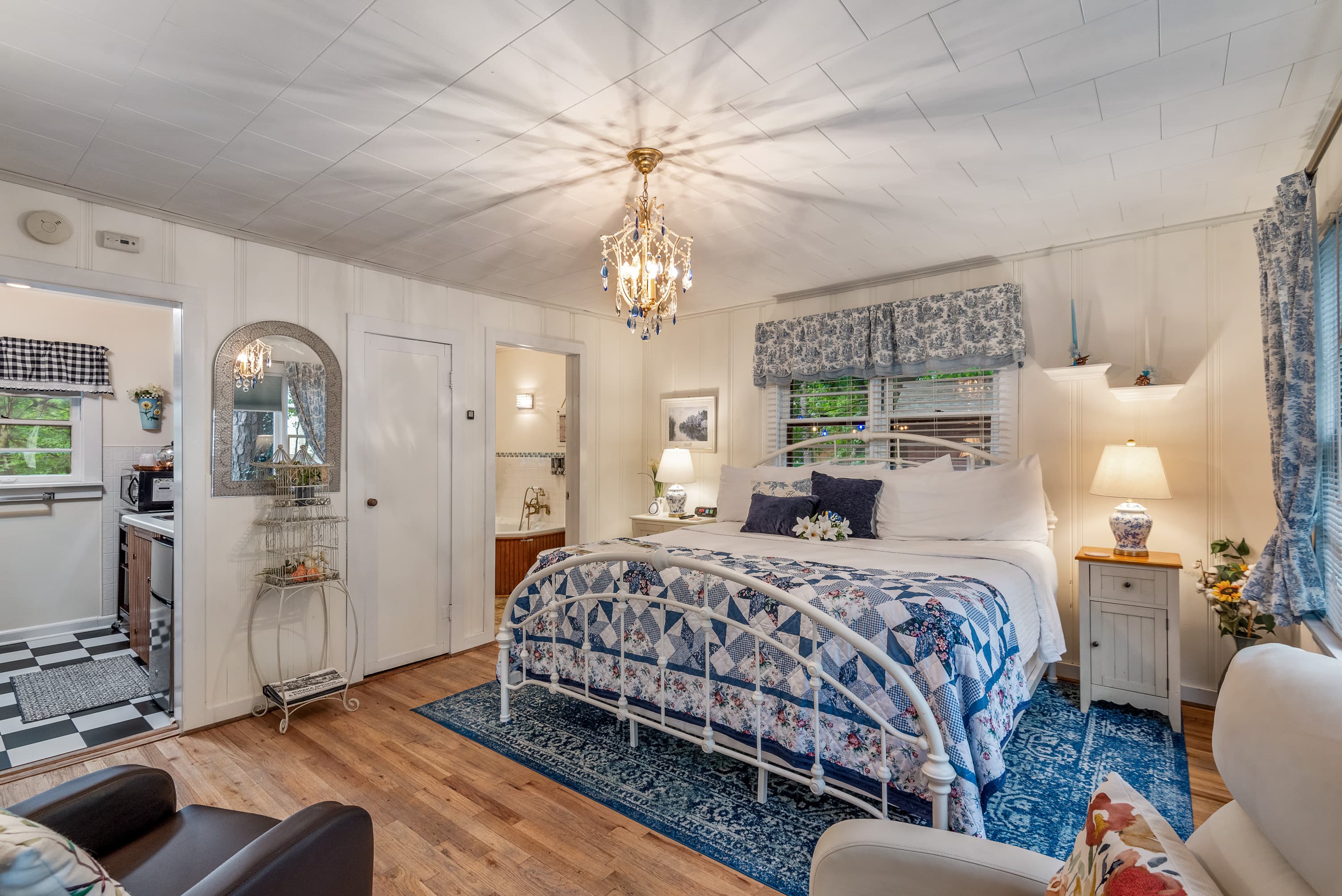 A spacious bedroom features a king-size bed with a white metal frame and blue-and-white bedding. The room has a small kitchenette, a crystal chandelier, and light-colored walls.