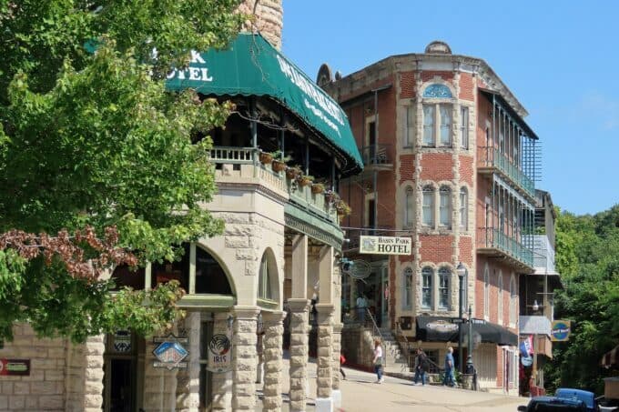 Two historic hotels stand side by side on a picturesque street, with vibrant greenery and clear blue skies above.