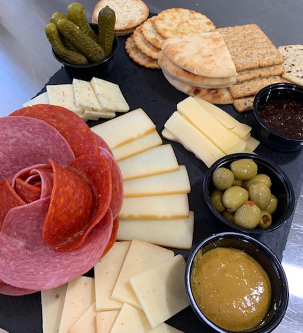 A charcuterie board featuring various cheeses, meats, olives, pickles, crackers, and dips arranged on a black slate.