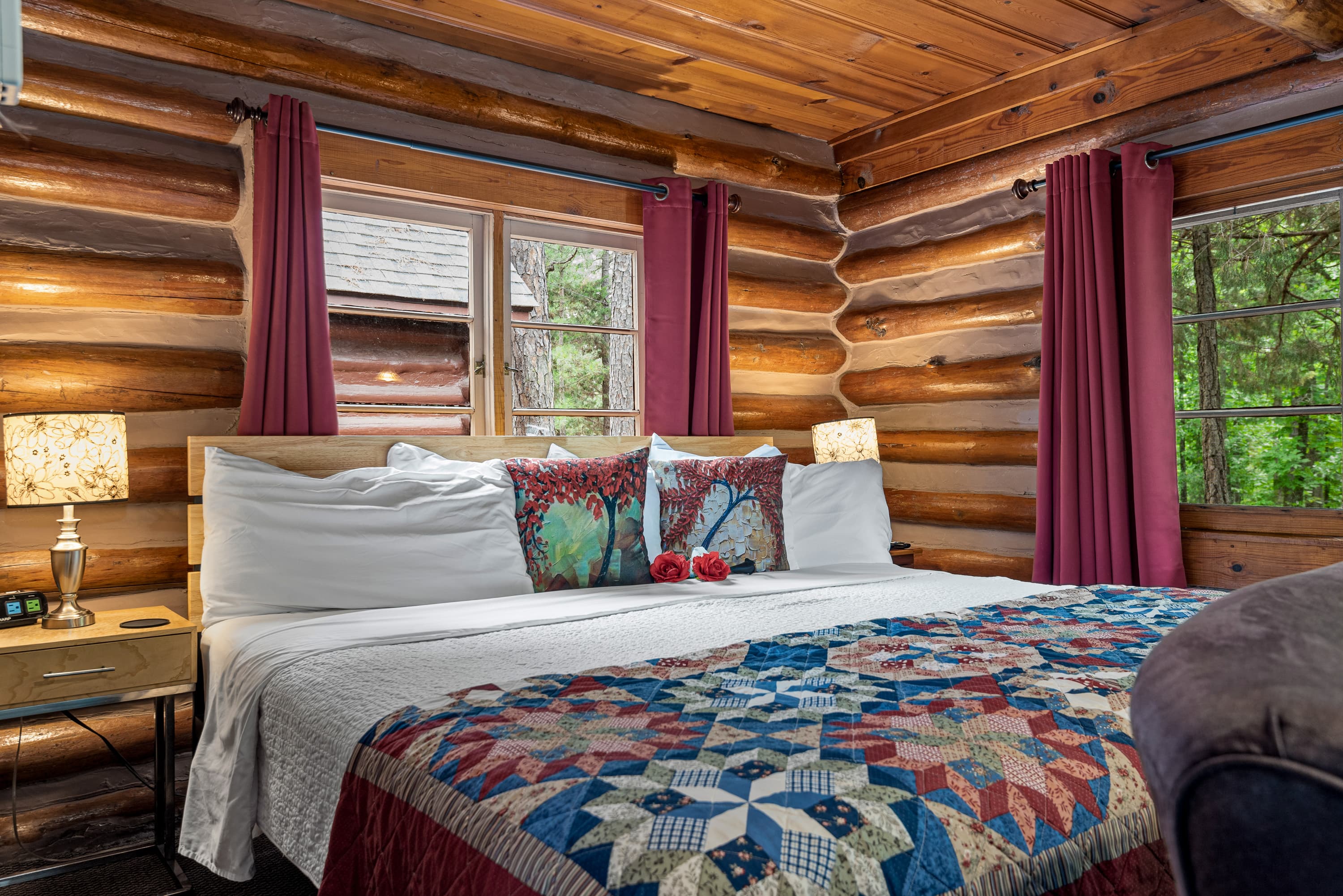 A cozy log cabin bedroom with a king bed, a colorful patchwork quilt, and deep red curtains. The room is decorated in a rustic style.