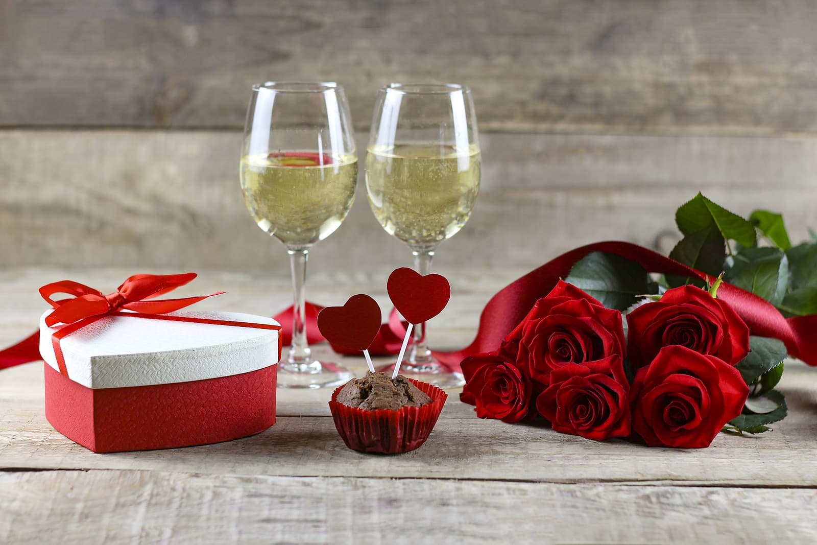 A romantic setup featuring two glasses of champagne, a red ribbon heart-shaped gift box, a chocolate cupcake with heart toppers, and a bouquet of red roses. A romantic setup featuring two glasses of champagne, a red ribbon heart-shaped gift box, a chocolate cupcake with heart toppers, and a bouquet of red roses.