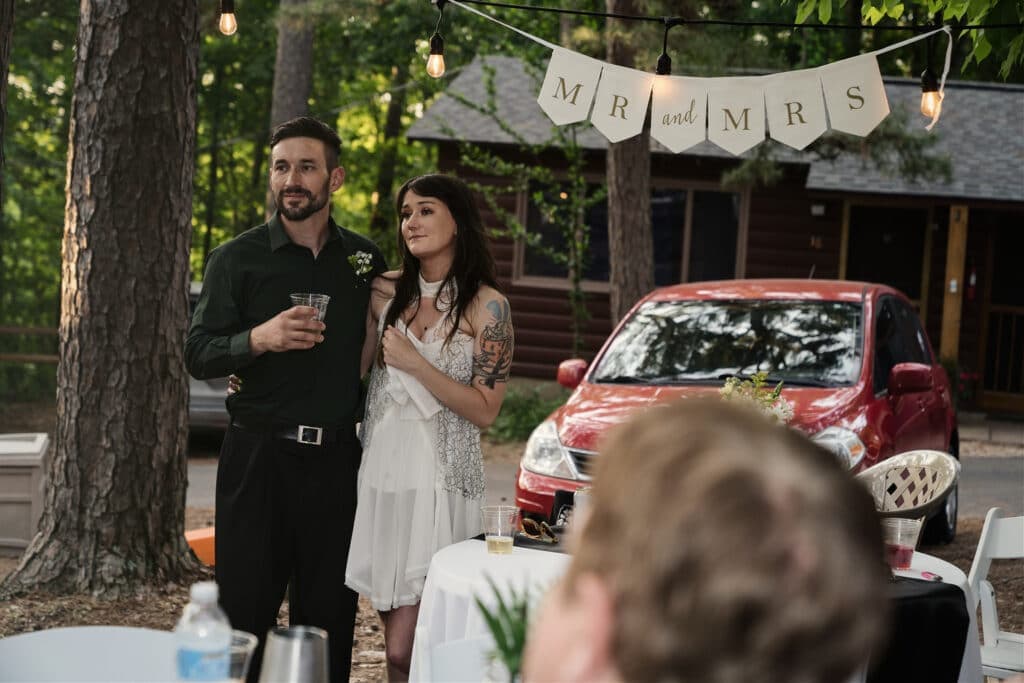 A couple stands together, holding drinks, under a banner reading "MR and MRS" at an outdoor celebration.