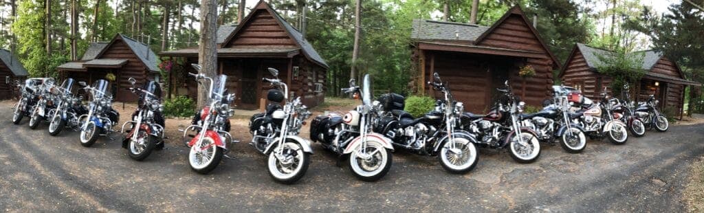 A row of various motorcycles parked in front of wooden cabins surrounded by trees.