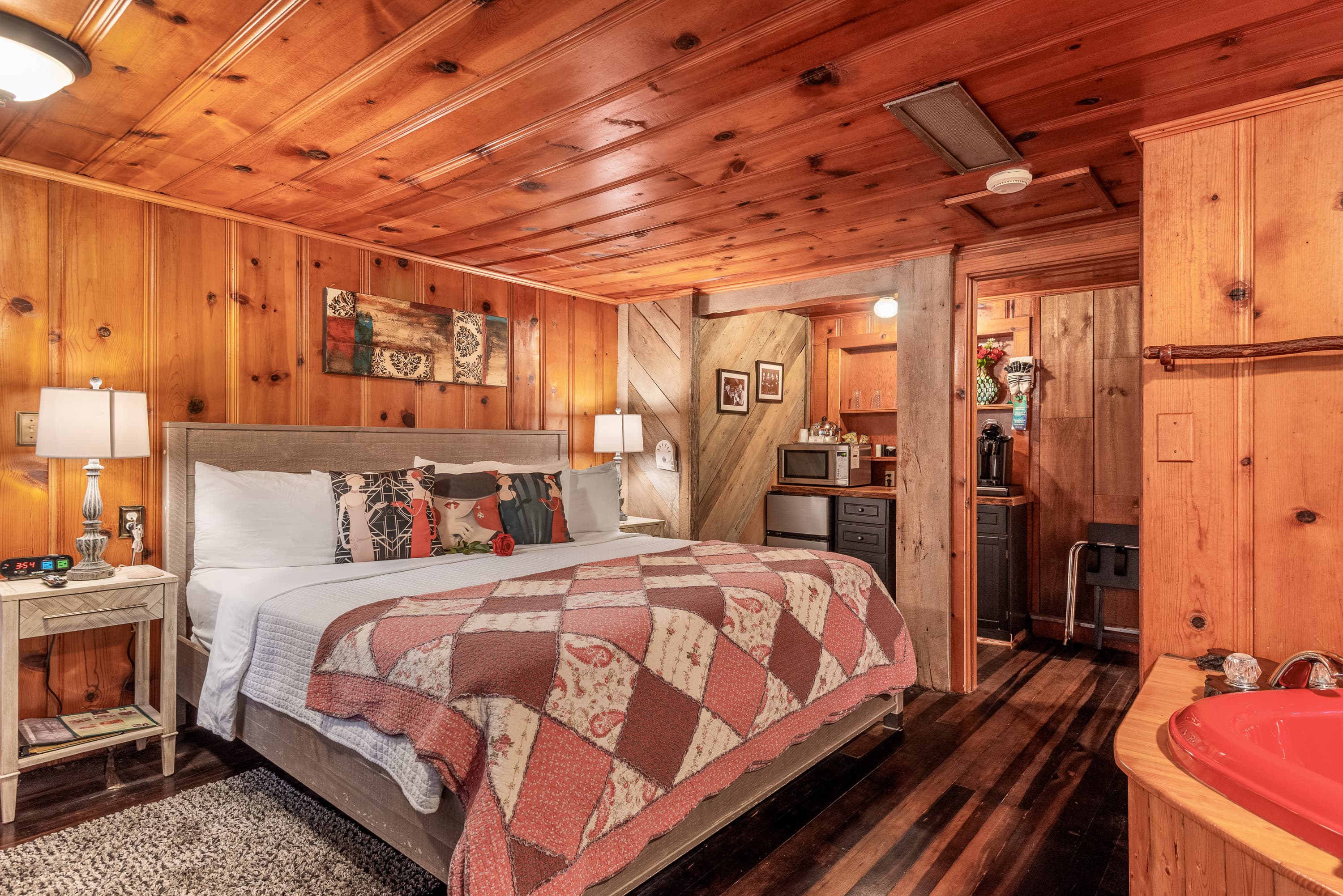 A spacious, rustic bedroom with wood-paneled walls and a king-size bed with a patchwork quilt. The room also features a red, heart-shaped jetted tub in the corner.
