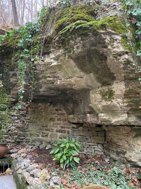 A moss-covered rock formation with a stone wall and greenery at its base.
