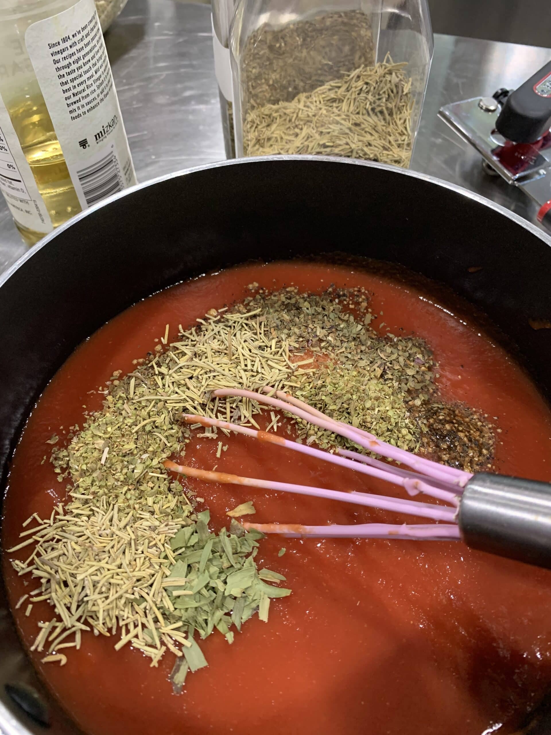 A whisk mixes herbs into a pot of tomato sauce on a stainless steel countertop.