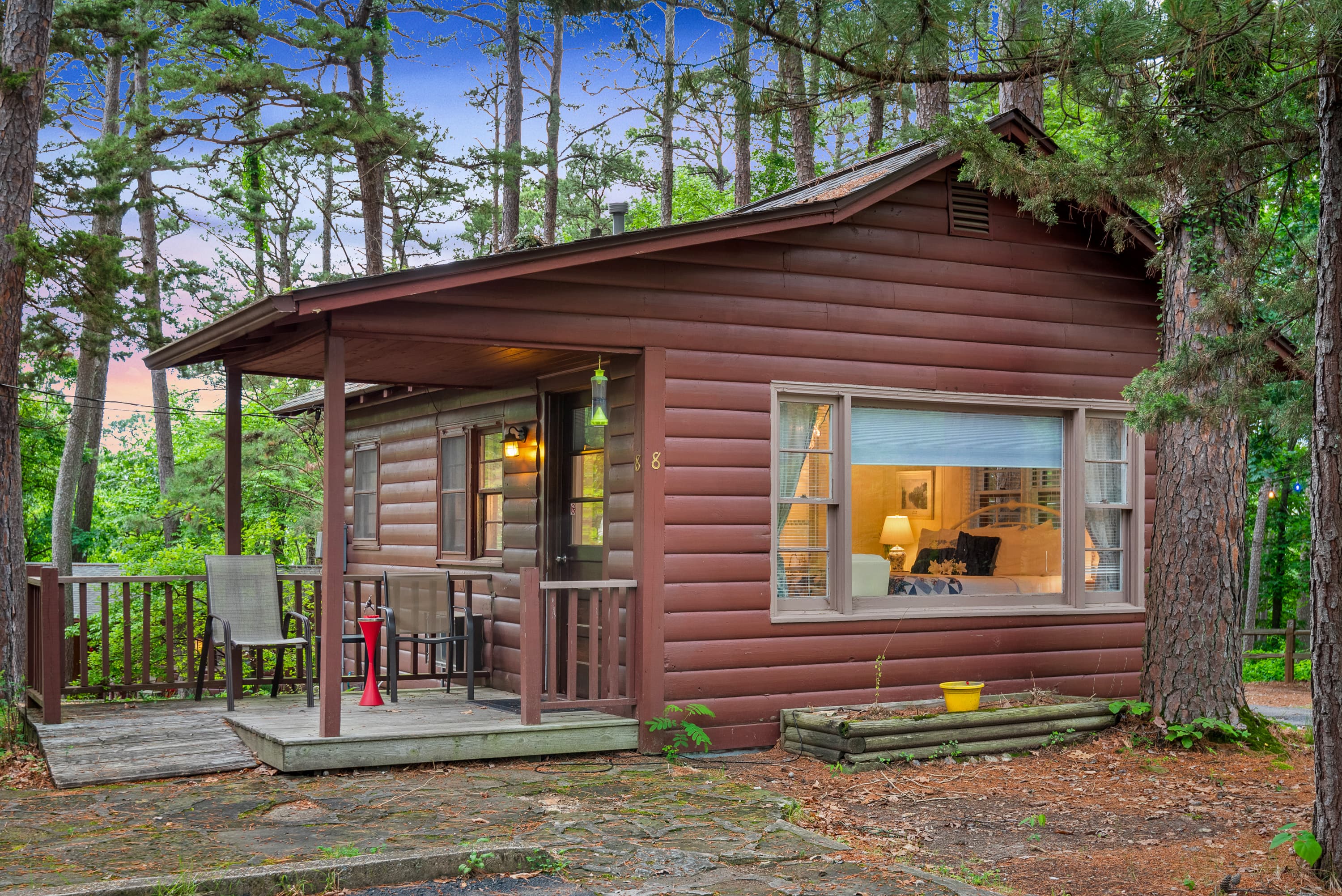 A charming, rustic duplex cabin with a small porch with a table and chairs. The cabin is nestled in a wooded area with tall pine trees.