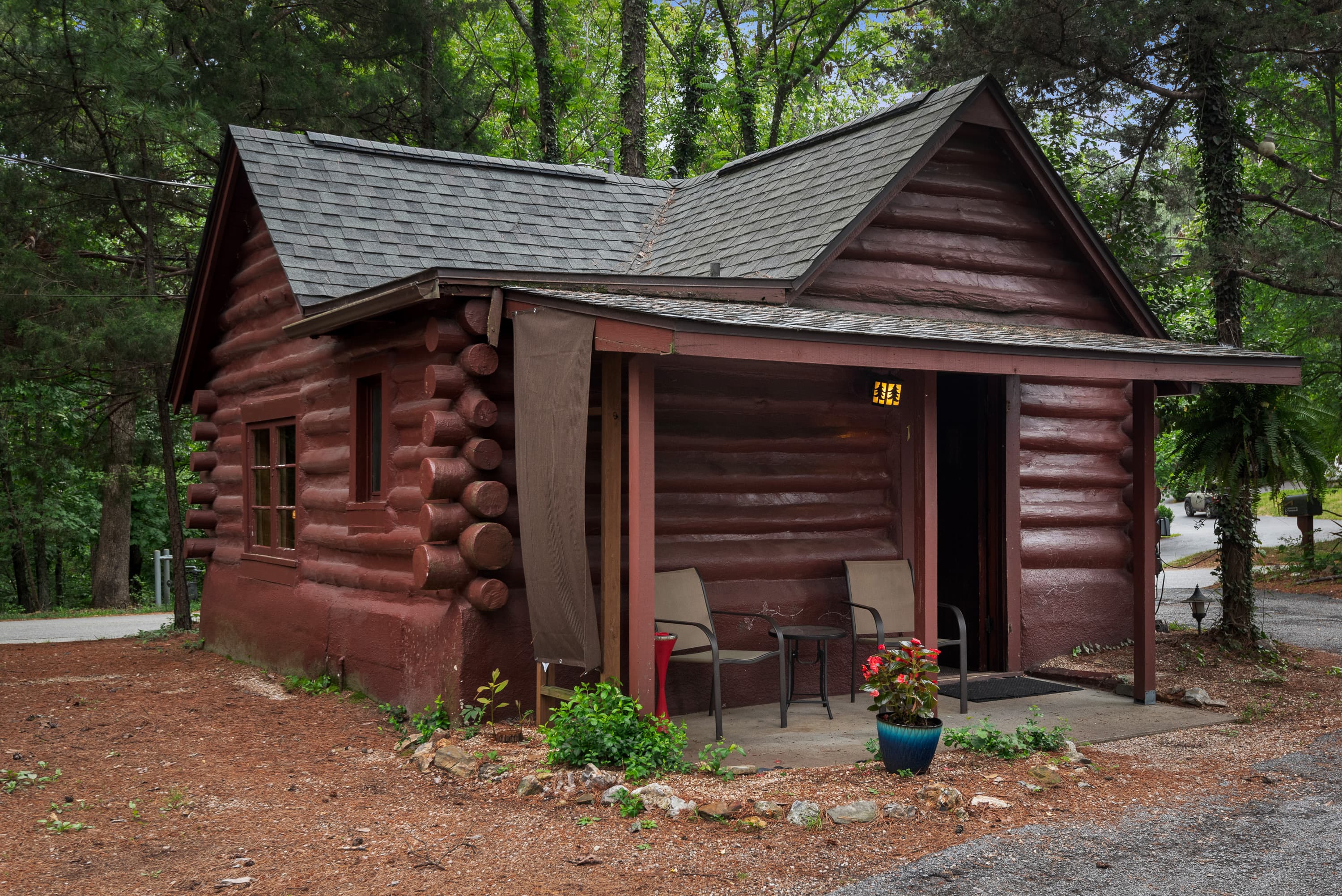 A charming log cabin is nestled among trees, with a covered porch holding two chairs and a small table. A small pot of flowers sits nearby.