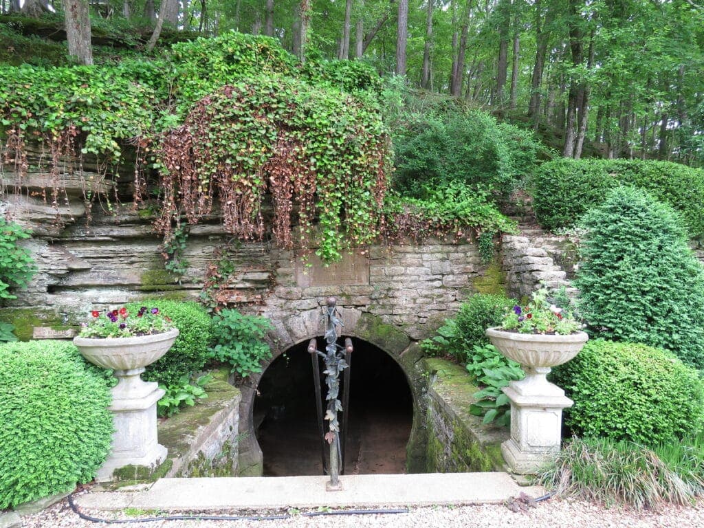 A stone archway partially covered in greenery leads into a dark tunnel, flanked by decorative urns filled with flowers.