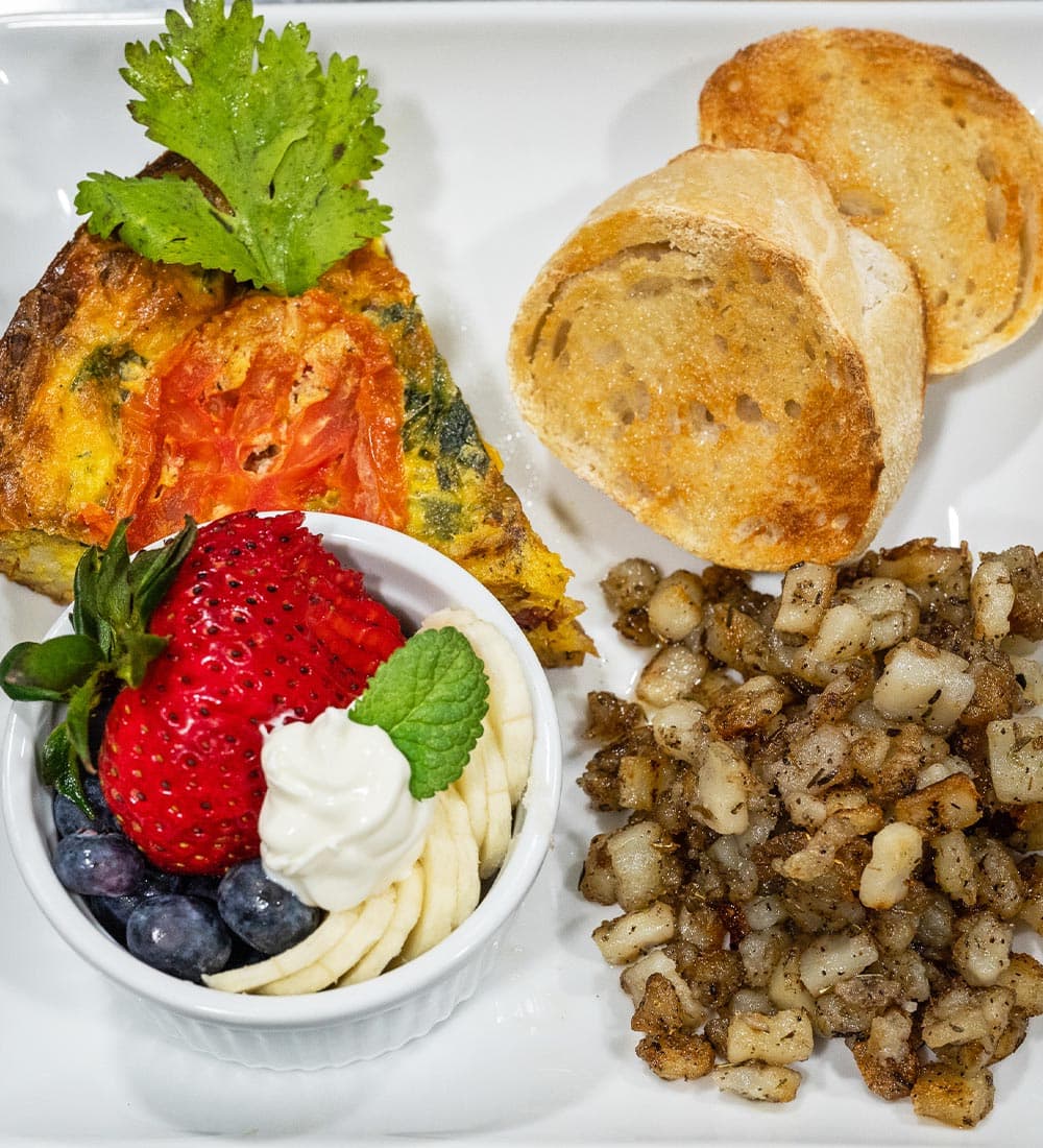 A plate featuring a slice of frittata with tomatoes, two pieces of toasted bread, a small bowl of mixed berries, and seasoned diced potatoes.