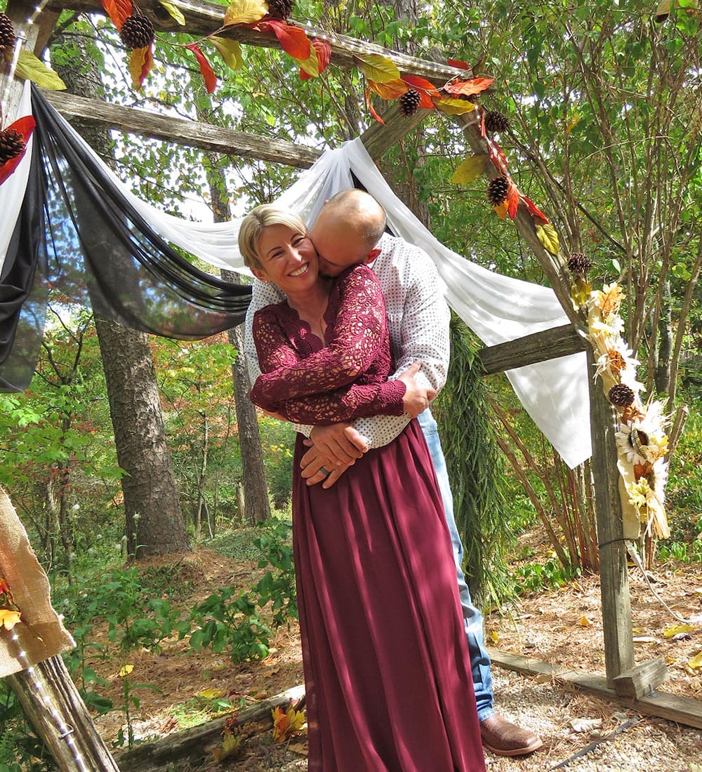 A couple embraces under a decorated archway in a wooded setting.