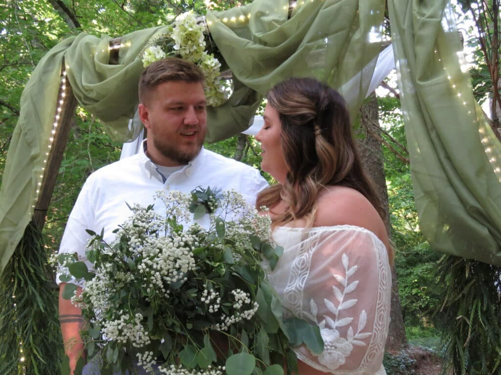 A couple stands together under a floral arch, exchanging loving glances during their outdoor wedding ceremony.