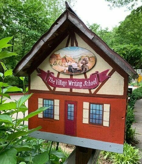 A decorative sign featuring "The Village Writing School" with a picturesque image above it, set against a lush green backdrop.