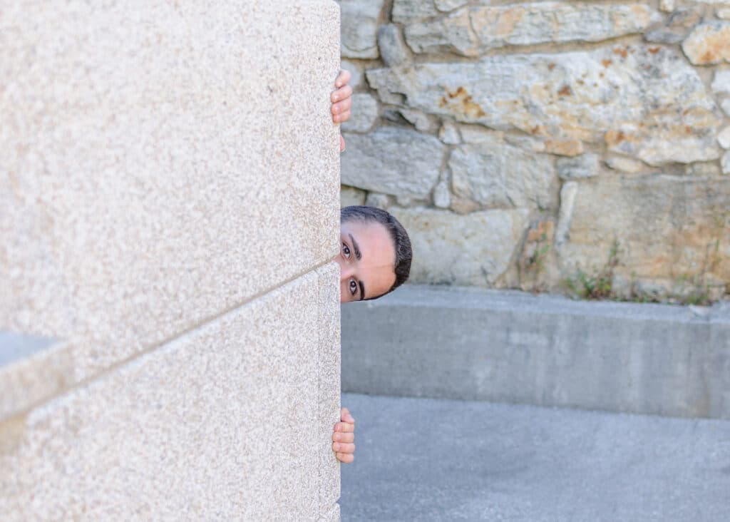 A person peeks around a concrete structure against a stone wall backdrop.