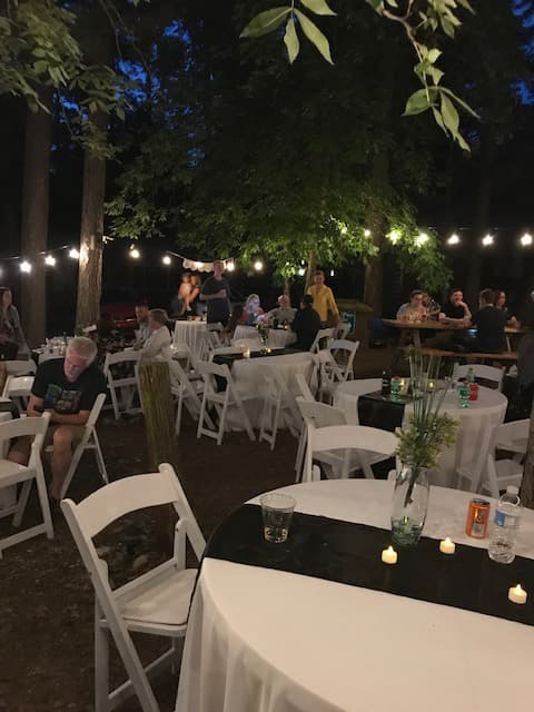 An outdoor gathering scene with tables and string lights set up in a wooded area during the evening.