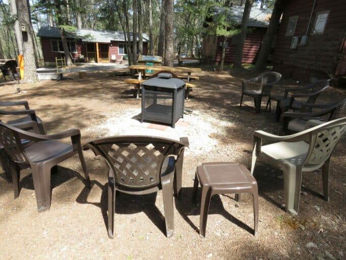 A circle of plastic chairs surrounds a fire pit in a wooded area with cabins in the background.