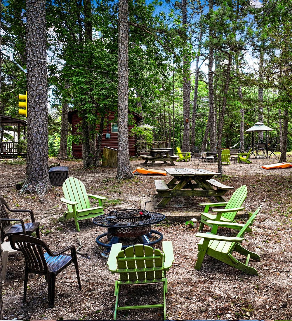 A cozy outdoor seating area surrounded by trees, featuring green and black chairs, fire pit, and picnic tables.
