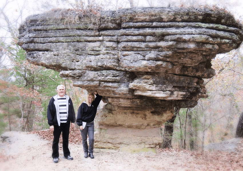 Two people stand under a large rock formation in a wooded area.