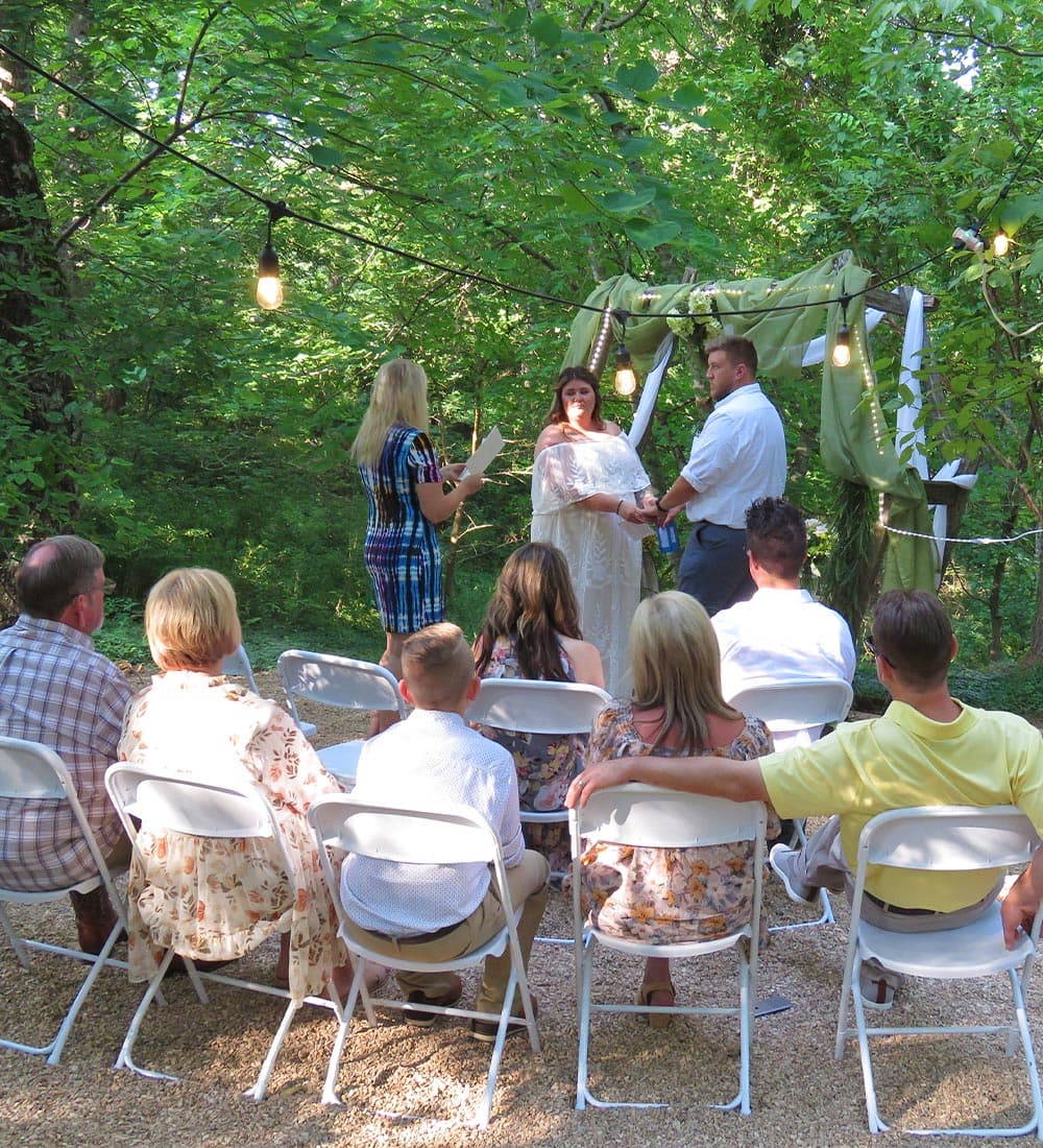 A couple exchanges vows at an outdoor wedding ceremony surrounded by guests in a lush green setting.