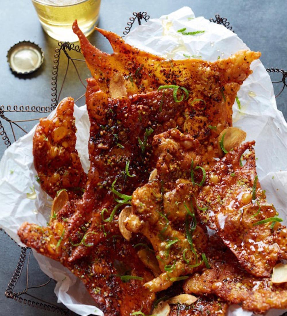 A close-up of crispy, seasoned pork cracklings garnished with green onion, served in a decorative basket alongside a glass of beer.