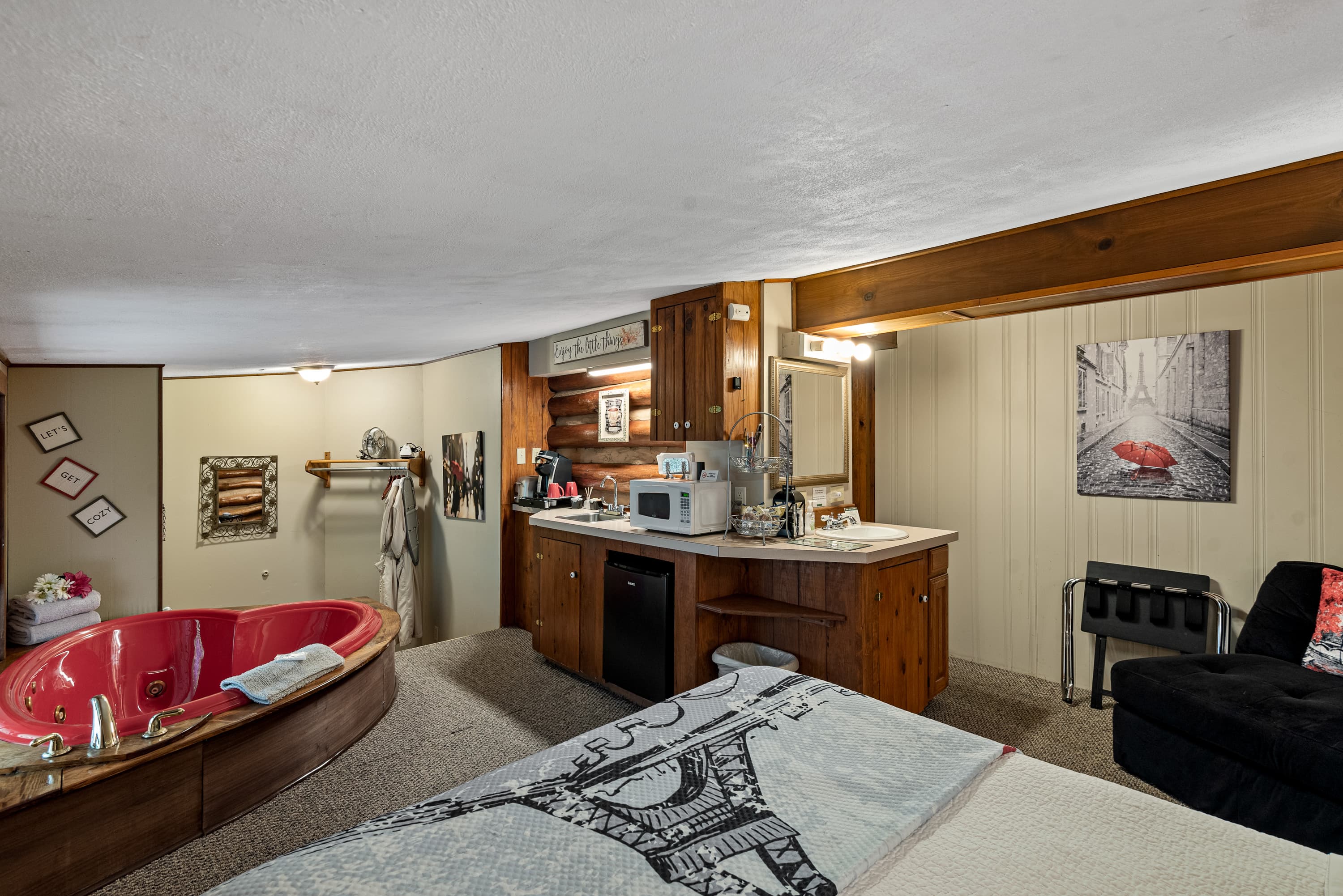 Cozy Hideaway features a red heart-shaped jetted tub, a small kitchenette, and a large bed with a blanket with a black and white image of the Eiffel Tower.